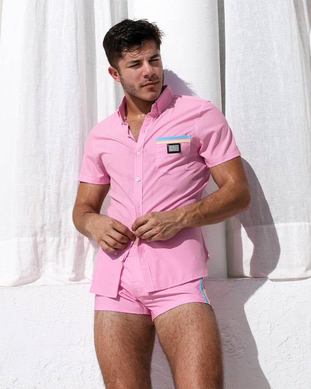 A muscular man models the PADAM PINK Stretch Shirt and matching Mini Shorts, standing against a white wall and sheer curtains in bright sunlight. The shorts feature colorful side stripes, adding a playful touch to the coordinated set. Designed by DC2, a premier men&