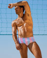 A man models THE KEN (IBIZA EDITION) Swim Sunga on a sunny beach, with a beach volleyball net and bright blue sky in the background. Created by BANG! Miami, a leading men's beachwear brand from Miami.