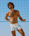 A man models THE KEN (MYKONOS EDITION) Beach Shorts in front of a beach volleyball net, showcasing the light blue and white striped design. His athletic build and the clear blue sky highlight the shorts' modern and stylish appeal. Designed by BANG! Clothes, a premier men's beachwear brand from Miami.