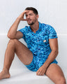 A man models the WET Stretch Shirt and matching beach shorts, sitting against a white textured wall. The vibrant blue outfit features a water-inspired pattern, designed for both comfort and fashion, perfect for resort or beachwear. Designed by DC2 Miami, a premier men's beachwear brand from Miami.