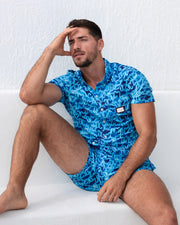 A man models the WET Stretch Shirt and matching beach shorts, sitting against a white textured wall. The vibrant blue outfit features a water-inspired pattern, designed for both comfort and fashion, perfect for resort or beachwear. Designed by DC2 Miami, a premier men's beachwear brand from Miami.