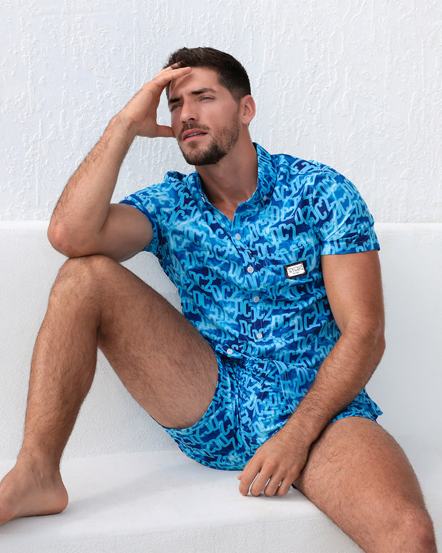 A man models the WET Stretch Shirt and matching beach shorts, sitting against a white textured wall. The vibrant blue outfit features a water-inspired pattern, designed for both comfort and fashion, perfect for resort or beachwear. Designed by DC2 Miami, a premier men&