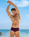 A man models the BERRY GOOD bordeaux plum wine Brazilian swim sunga on a sunny beach. The vibrant blue sky and ocean backdrop brings out the swimwear's trendy look. Designed by DC2, a premier men's beachwear brand from Miami.