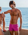 A male model wearing CONFESS MAGENTA Mini Shorts by Bang Clothes standing by the ocean. The bright magenta shorts highlight his fit physique, with the calm sea and a clear sky in the background.