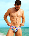 Male model adjusting colorful MOSAIC swim briefs by BANG! Clothing, featuring vibrant geometric patterns. These form-fitting men's swim briefs offer a bold, unique design perfect for summer beachwear and poolside style.