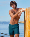 Model leaning against a wooden post by the ocean, wearing DC2 Miami MONO TEAL Tailored swimming Shorts. These men's beach shorts feature a rich teal color with a subtle geometric pattern and adjustable side buckles, offering a tailored fit perfect for a rich luxury beach or poolside look.