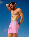 Model wearing DC2 Miami PADAM PINK Swim Shorts, featuring a soft pink fabric with colorful side stripes. These tailored swim shorts offer a relaxed fit with a drawstring waist, perfect for casual beachwear or poolside lounging under Miami's clear blue sky.