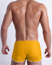 Back view of male model wearing the AMBER SAND beach Swim Shorts for men by BANG! Miami in a solid dark yellow color.