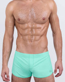 Model wearing AQUA LITE swim shorts in a crisp seafoam green shade. These square-cut swim bottoms offer a perfect mid-thigh length with a smooth, body-contouring fit designed for elevated summer vibes and confident style.