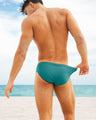 Male model wearing ATLANTIS TEAL Swim Briefs by DC2 Miami, adjusting the waistband while standing on a sandy beach. These vibrant teal men's swim briefs offer a comfortable and stylish fit, perfect for beachwear and summer relaxation.