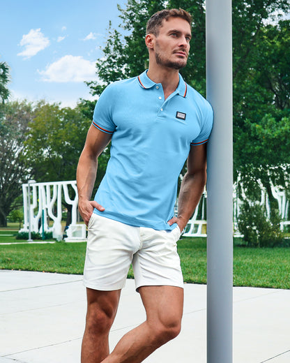 Model wearing DC2 Biscayne Blue Pima Polo Shirt and White Lotus Street Shorts, standing outdoors in a modern park setting, showcasing a crisp, polished resort look.