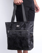 Side view of the BLACK CAMO Poolside Tote Bag Foldable/Portable by DC2 brand. Features smooth zipper in the front and the side, water resistant fabric, and the DC2 logo in a metallic silver plaque.