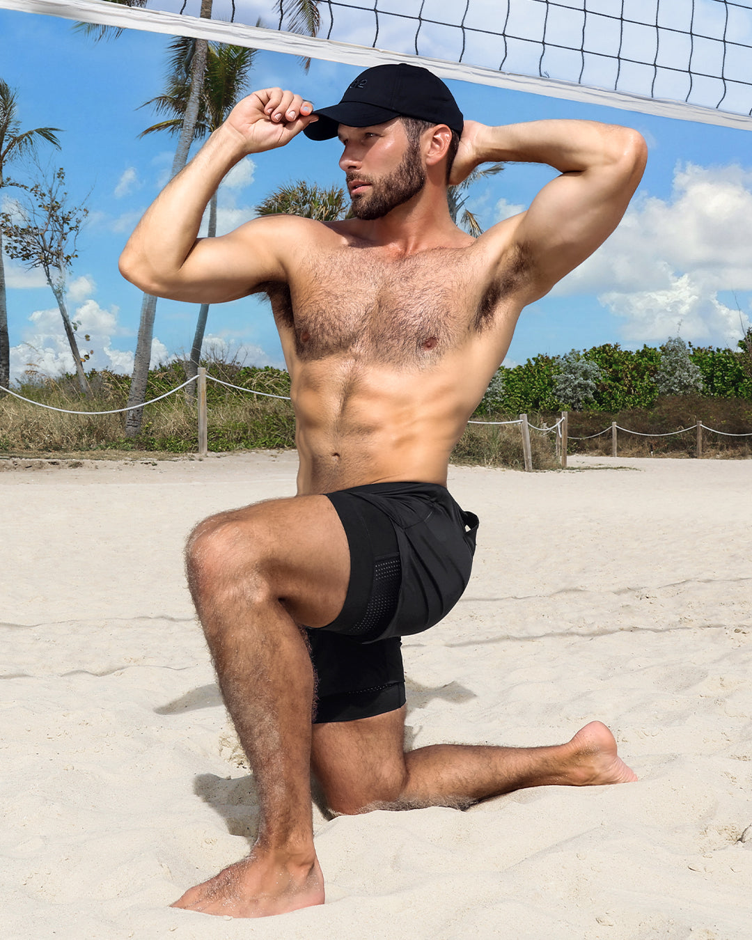 Model wearing DC2 Black Endurance 2-in-1 Workout Shorts, kneeling on the sand at a beach volleyball court, showcasing the athletic fit and breathable mesh side panels.