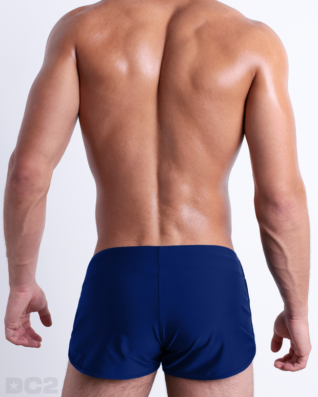 Back view of male model wearing the BLUE BY THE OCEAN beach Swim Shorts for men by BANG! Miami in a solid dark blue color.