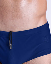 Close-up view of the BLUE BY THE OCEAN men’s drawstring briefs showing white cord with custom branded metallic silver cord ends, and matching custom eyelet trims in silver.