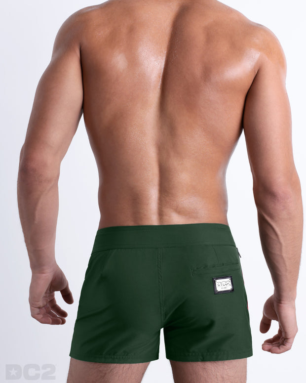 Back view of a male model wearing men’s Summer BRAVE GREEN Beach Shorts in a solid dark green color with orange and white side stripes, complete the back pockets, made by DC2 a capsule brand by BANG! Clothes in Miami.