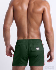 Back view of a male model wearing men’s BRAVE GREEN Flex Shorts swimsuits in a solid green color with white and orange side stripes, complete the back pockets, made by DC2 a capsule brand by BANG! Clothes in Miami.