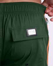 Close-up view of the BRAVE GREEN men’s Flex Shorts back pocket, showing custom branded silver metal logo.