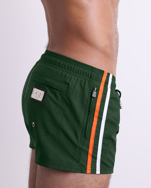 Side view of the BRAVE GREEN for men’s summer Poolside Shorts with dual zippered pockets. The shorts are in a solid green color with side orange and white stripes for men made by DC2 a brand based in Miami.