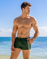 Male model wearing BRAVE GREEN Poolside Shorts by DC2 Miami, standing by the beach with a relaxed pose. These deep green men's swim shorts feature stylish white and orange racing stripes and a drawstring waistband, ideal for poolside lounging and beach activities.