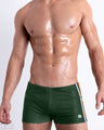 Front view of model wearing the BRAVE GREEN men’s Swim Trunks, premium swimwear in a solid dark green color with white and orange stripes on the sides for men. These high-quality swimwear are by DC2, a men’s beachwear brand from Miami.