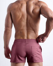 Back view of male model wearing the BUST A MAUVE beach trunks in a light mauve color for men by BANG! Miami.