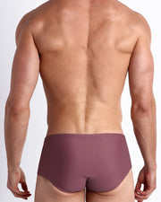 Back view of a male model wearing men’s BUST A MAUVE Brazilian Sunga swimwear in a light mauve purple color made with Italian-made Vita By Carvico Econyl Nylon by the Bang! Clothes brand of men's beachwear.