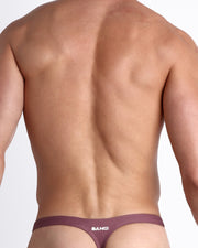 Back view of a male model wearing men’s BUST A MAUVE swim thong in a light purple wine color made with Italian-made Vita By Carvico Econyl Nylon with official logo of BANG! Brand in white.