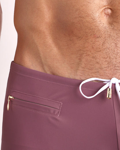 Close-up view of the BUST A MAUVE Swim Trunks mens swimsuit in a mauve color with mini zippered pocket and white internal drawstring cord showing custom branded golden buttons by BANG! clothing brand.