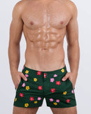 Frontal view of a masculine model wearing the BUY MYSELF FLOWERS beach shorts for men by DC2 Miami, a premium swimwear brand. These modern-fit swimming shorts feature a deep green base with vibrant red, pink, and yellow floral prints, offering a bold, stylish look perfect for beach days, poolside lounging, and summer adventures.