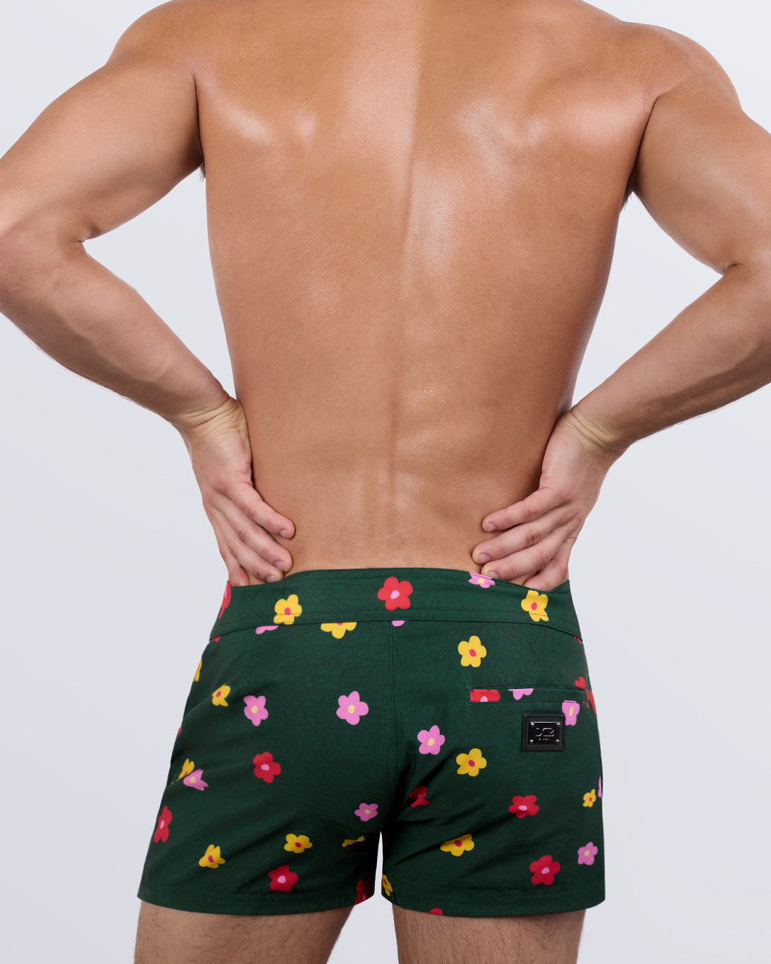 Back view of a male model wearing the men's BUY MYSELF FLOWERS beach shorts by DC2 Miami. Featuring a playful floral pattern on a rich green backdrop, a sleek fit, and a functional back pocket with a DC2 signature metal logo plaque, these premium swim shorts combine eye-catching design with ultimate comfort.
