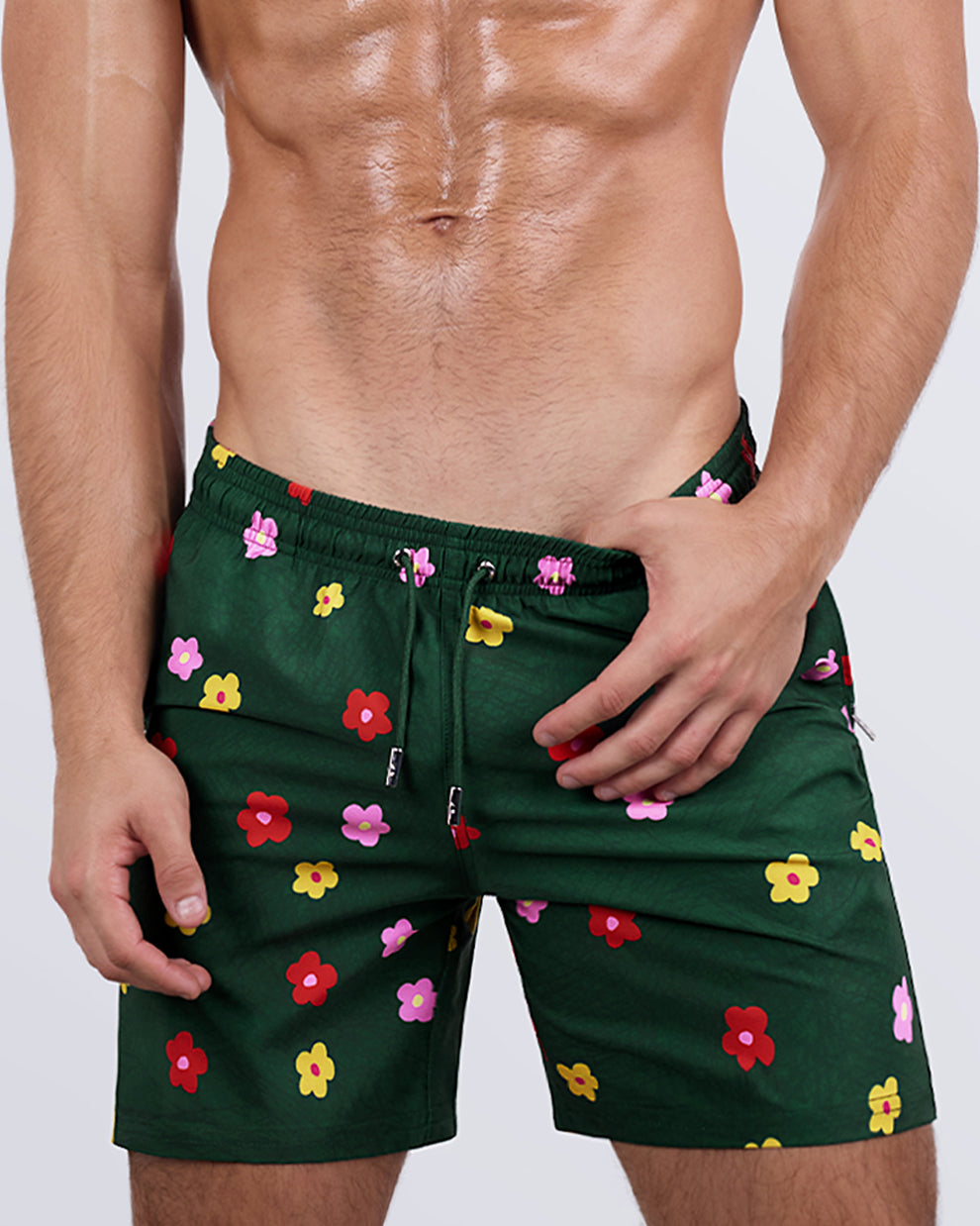 Frontal view of a male model wearing the BUY MYSELF FLOWERS Resort Shorts for men by DC2 Miami. These stylish dark green swim shorts feature a playful, all-over floral print in red, yellow, and pink, an elastic waistband with an adjustable drawstring, and a modern above-the-knee cut for a fresh, confident summer look.