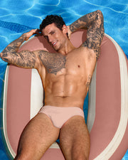 Male model posing in a pool wearing the NAKED PINK men’s European Swim Brief bikini in a light pink color with official logo of BANG! Brand in white by the Bang! Menswear brand from Miami.