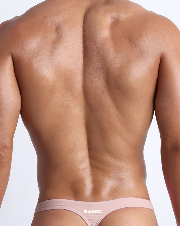 Back view of a male model wearing men’s NAKED PINK swim thong in a solid light pink color with official logo of BANG! Brand in white.