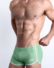 Side view of a masculine model wearing men’s swimwear in a solid green color made with Italian-made Vita By Carvico Econyl Nylon with official logo of BANG! Brand in white.