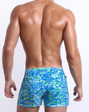 Male model wearing men’s CAMO POP (BLUE/GREEN) Beach Shorts swimsuit in a colorful blue and green neon camo print, complete with a back pocket, designed by BANG! Clothes in Miami.