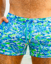 Male model lying on the sand wearing CAMO POP (Blue/Green) Show Shorts by BANG! Clothing. These vibrant camouflage-patterned men's beach shorts are perfect for summer beachwear and tropical vacations.