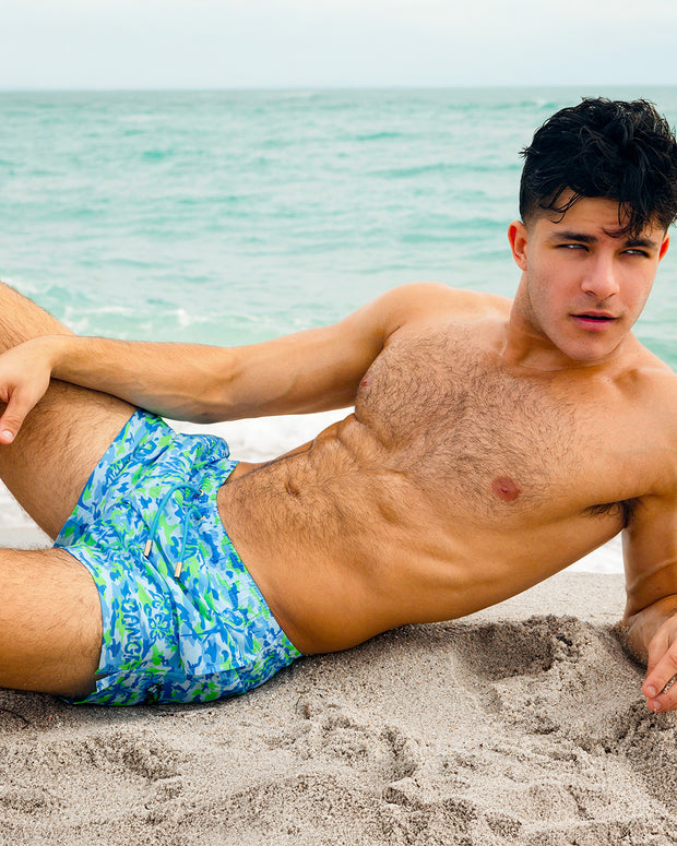 At the beach, a male model is wearing the CAMO POP (BLUE/GREEN) Show Shorts. The swimsuit features a bright sky blue and green neon camo print designed by BANG! Clothes in Miami.