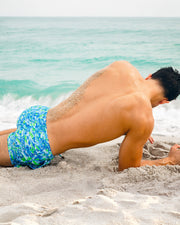 Male model lying on the sand wearing CAMO POP (Blue/Green) Show Shorts by BANG! Clothing. These vibrant camouflage-patterned men's beach shorts are perfect for summer beachwear and tropical vacations.