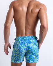 Male model wearing men’s CAMO POP (BLUE/GREEN) Tailored Shorts swimsuit in a colorful blue and green neon camo print, complete with a back pocket, designed by BANG! Clothes in Miami.