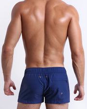 Back view of male model wearing men’s CASINO ROYALE (BLUE) beach Resort Shorts swimsuits in a blue color. Inspired by actor Daniel Craig's iconic blue swim trunks worn in the 2006 film Casino Royale, complete with a zippered back pocket, are designed by BANG! Clothes in Miami.