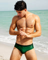 Model wearing CASINO ROYALE (GREEN) men's swim briefs by BANG! Clothing on a beach. Premium men's swimwear featuring a sleek fit and high-quality fabric, perfect for summer beach fashion and Miami beachwear. Stylish and comfortable swim briefs designed for active beach lifestyles.