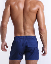 Back view of male model wearing men’s CASINO ROYALE (BLUE) shorter length swimming Flex Shorts in a blue color, complete with a back pocket. Inspired by actor Daniel Craig's iconic blue swim trunks worn in the 2006 film Casino Royale, are designed by BANG! Clothes in Miami.