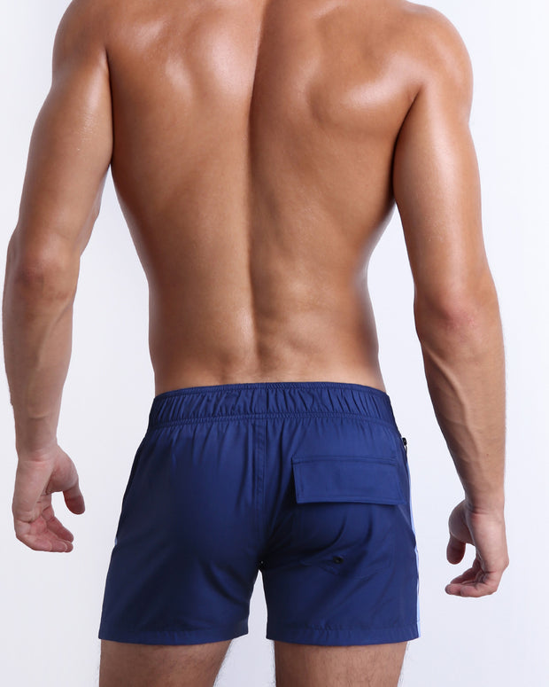 Back view of male model wearing men’s CASINO ROYALE (BLUE) shorter length swimming Flex Shorts in a blue color, complete with a back pocket. Inspired by actor Daniel Craig&