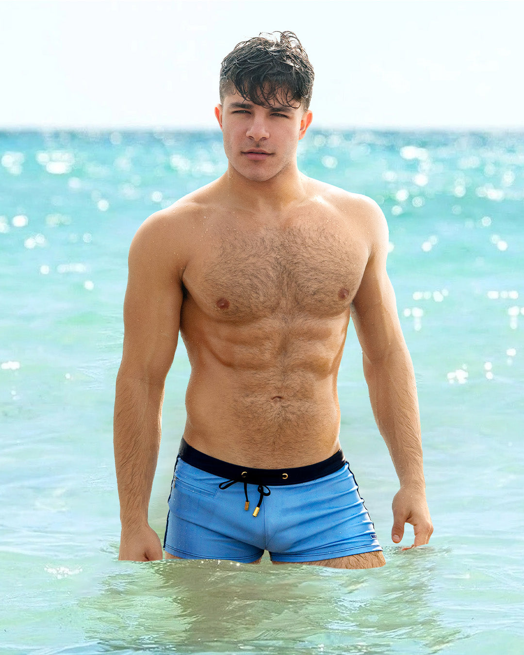 Male model at the beach wearing the CASINO ROYALE (BLUE) Swim Trunks. This Summer square-cut swim trunk swimsuit for the beach with a stylish color block design in dark blue for men. This high-quality swimwear item is by BANG! Clothes, a men’s beachwear brand from Miami. Inspired by the iconic style of Daniel Craig's blue swim trunks in the 2006 film Casino Royale.