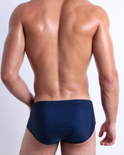 Back view of a model wearing CASINO ROYALE (BLUE) men’s beach sexy swim sunga in dark blue color. Inspired by actor Daniel Craig's iconic blue swim trunks worn in the 2006 film Casino Royale, is designed by BANG! Clothes in Miami.