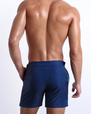 Back view of male model wearing men’s CASINO ROYALE (BLUE) long-length beach trunks in a blue color, complete with a back pocket. Inspired by actor Daniel Craig's iconic blue swim trunks worn in the 2006 film Casino Royale, are designed by BANG! Clothes in Miami.