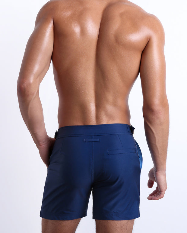 Back view of male model wearing men’s CASINO ROYALE (BLUE) long-length beach trunks in a blue color, complete with a back pocket. Inspired by actor Daniel Craig&