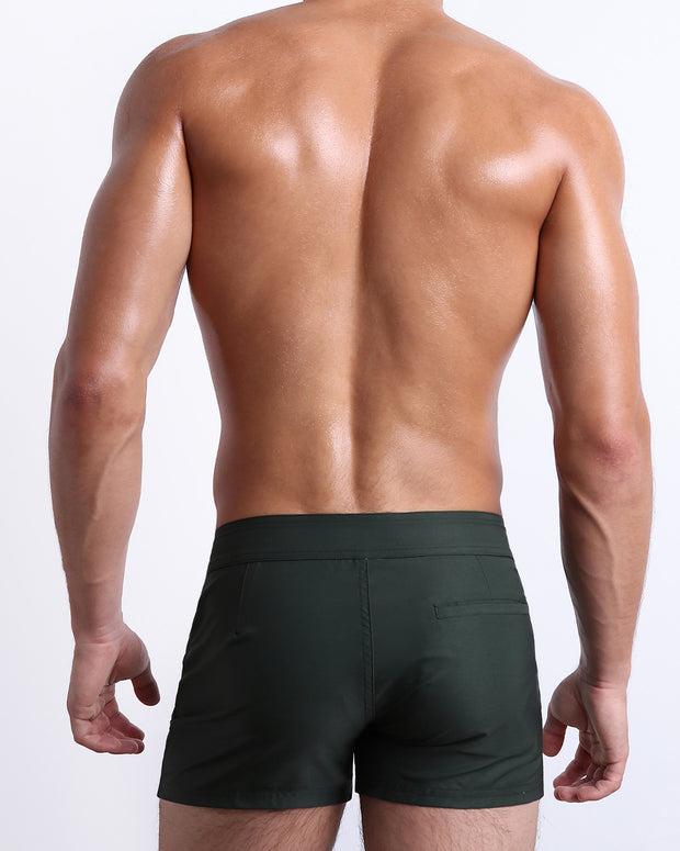 Back view of male model wearing men’s CASINO ROYALE (GREEN) beach Beach Shorts swimsuits in a blue color. Inspired by actor Daniel Craig&