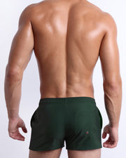 Back view of male model wearing men’s CASINO ROYALE (GREEN) beach Show Shorts swimsuits in a green color. Inspired by actor Daniel Craig's iconic blue swim trunks worn in the 2006 film Casino Royale, are designed by BANG! Clothes in Miami.